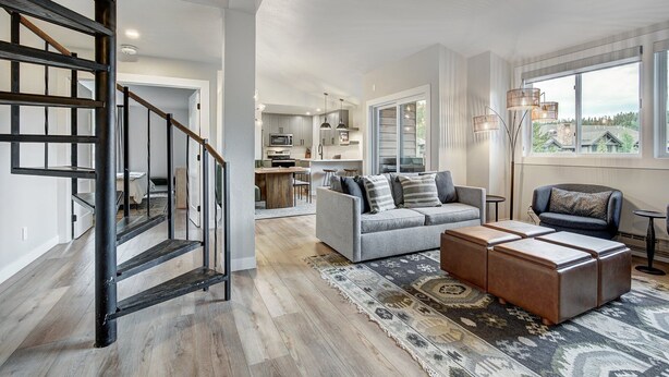 A spacious living area with a modern staircase, a grey sofa, a brown ottoman, and a view of the kitchen and outside.
