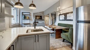 A modern kitchen with a central island, stainless steel appliances, pendant lighting, and a dining area with a green sofa and a wooden table.
