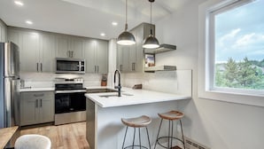 A modern kitchen with grey cabinets, a stainless steel refrigerator, a microwave, an oven, and a sink. There are two bar stools and a window providing a view of trees.