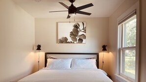 Un dormitorio con cama, ventilador de techo, ventana y una obra de arte colgada en la pared.