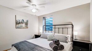 Une chambre à coucher avec un lit, une table de chevet, un ventilateur de plafond, une fenêtre donnant sur un beau paysage et un tableau accroché au mur.