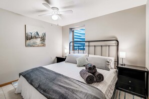 Un dormitorio con cama, mesita de noche, ventilador de techo, ventana con vistas y un cuadro en la pared.