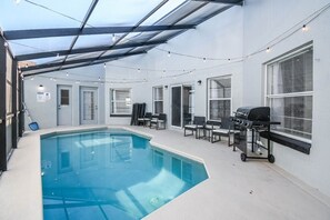 A covered outdoor swimming pool with a grill and chairs, surrounded by windows and a pergola. - Disney & Universal Studios Vacation Home (Kissimmee)