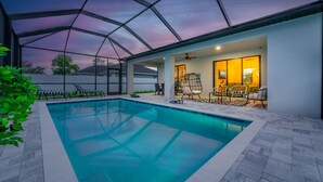 A covered swimming pool with lounge chairs and a ceiling fan, surrounded by a white wall and a view of the sky.