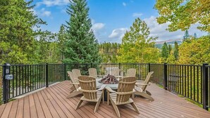Een houten terras met een vuurkorf en stoelen, omgeven door een zwarte balustrade, met uitzicht op een bebost gebied.
