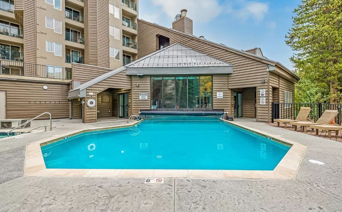 A swimming pool with a tiled deck, surrounded by a building with a sloped roof and large windows.