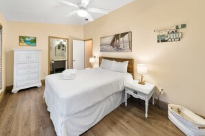 A hotel room with a large bed, a white dresser, a vanity with a mirror, a ceiling fan, and a beach-themed painting.