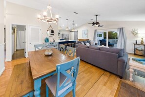 A spacious living area with a wooden dining table, blue chairs, a grey sofa, and a kitchen in the background.