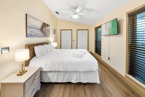 Ein Schlafzimmer mit einem Bett, Nachttischen, einem Deckenventilator, einem Gemälde, einem Fernseher und einem Fenster mit Jalousien.
