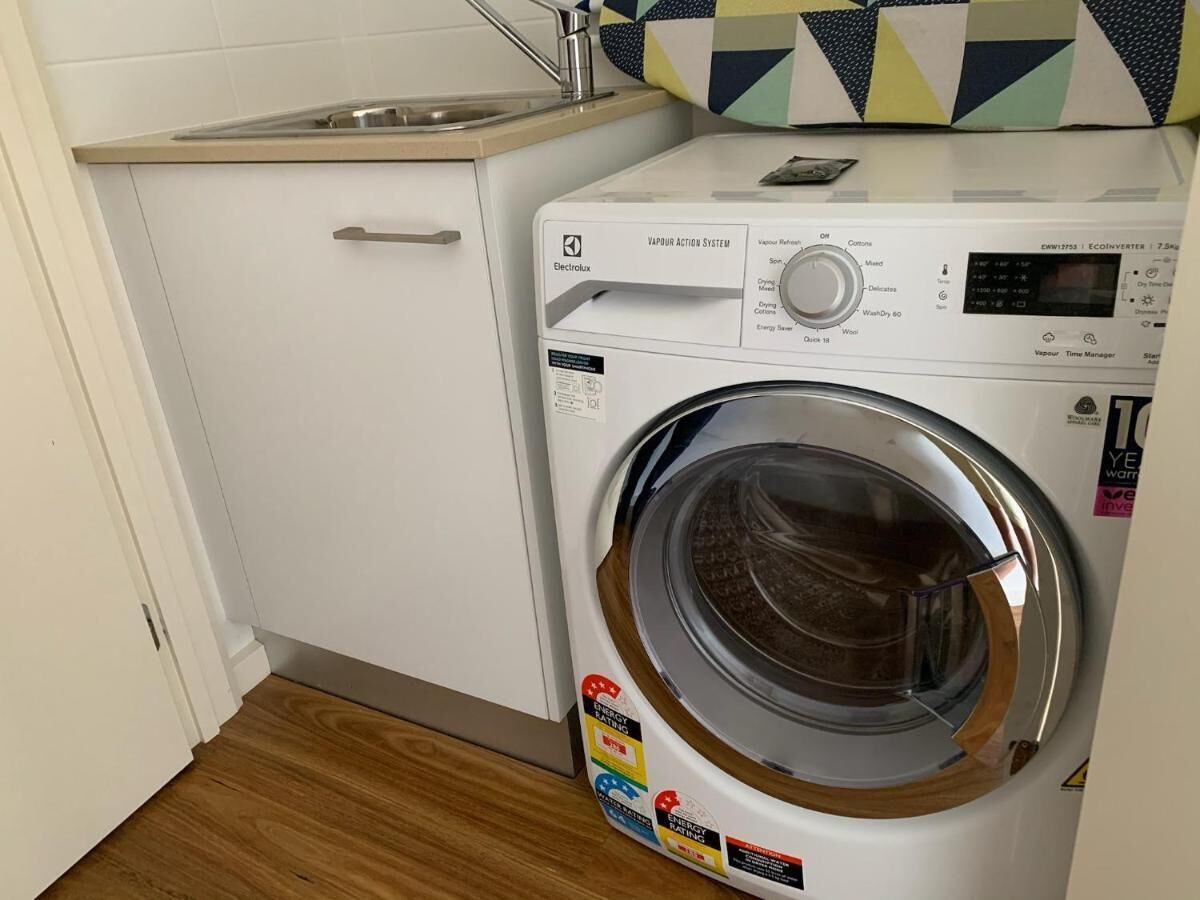 A compact, modern washing machine with a digital display, situated in a small laundry room with a wooden floor and a white cabinet.