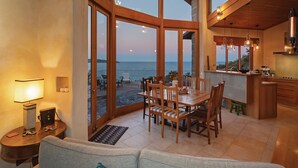 Een ruime woonkamer met een grote eettafel, houten stoelen en een bank. Een keuken is zichtbaar met een spoelbak en aanrecht. Grote ramen bieden uitzicht op de oceaan.