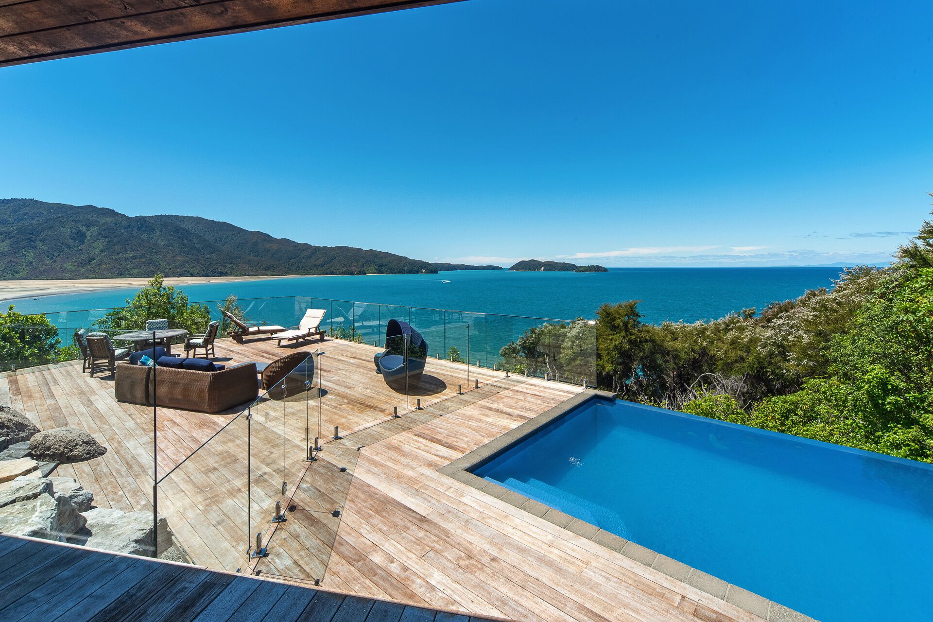 A wooden deck with a pool, lounge chairs, and a glass railing overlooking a beach and ocean.