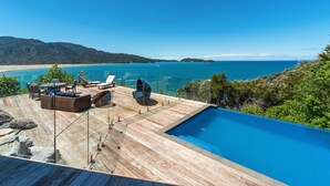 Een houten terras met een zwembad, lounge stoelen en een glazen balustrade met uitzicht op een strand en de oceaan.