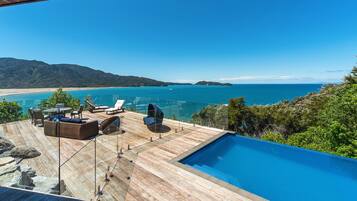 A wooden deck with a pool, lounge chairs, and a glass railing overlooking a beach and ocean.