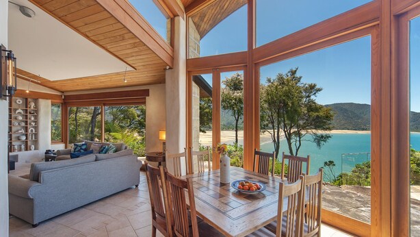 Een ruime woonkamer met een houten eettafel en stoelen, een bank en uitzicht op een strand en bergen door grote ramen.