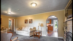 Una sala de estar con sofá, sillones, una mesita y una chimenea. Se ve una cocina a través de un arco.