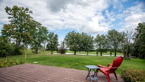 Una terraza de madera con una silla roja y una mesita pequeña, con vista a un área verde con árboles y un cielo despejado.