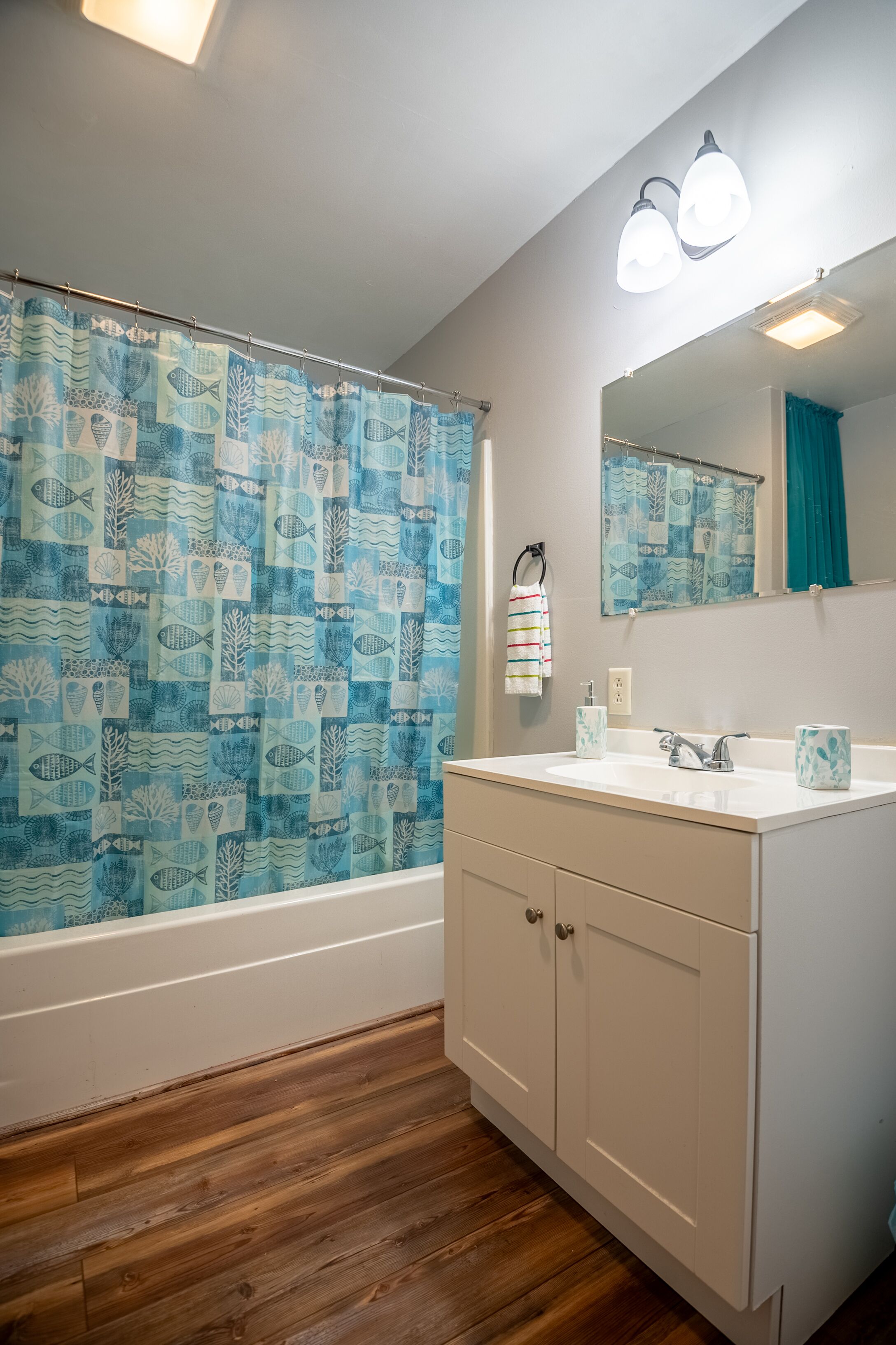 Une salle de bain avec un rideau de douche orné d’un motif de poissons bleus et blancs, un lavabo blanc avec un miroir au-dessus, et un plancher en bois.