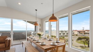 Un comedor con mesa de madera dispuesta para seis comensales, ventanas amplias con vista al océano e iluminación colgante.