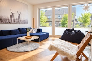 Ein modernes Wohnzimmer mit einem blauen Sofa, einem runden Couchtisch, einem weißen Teppich, einem gemütlichen Sessel und einem großen Fenster mit Blick auf Häuser und Bäume.