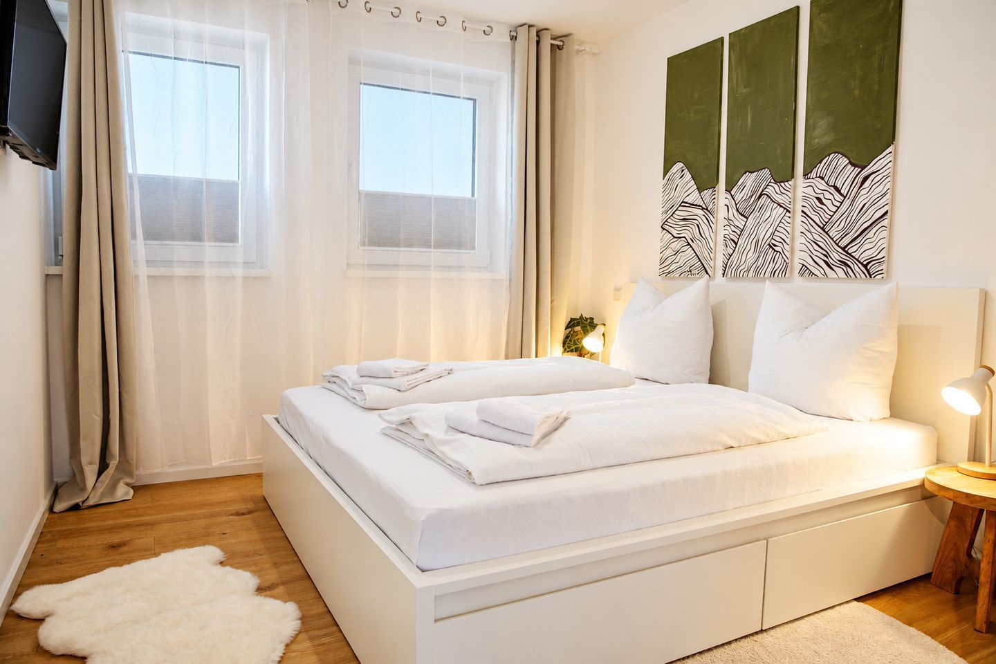 A modern bedroom with a large bed, white bedding, a small bedside table with a lamp, and a view of the outdoors through sheer curtains.
