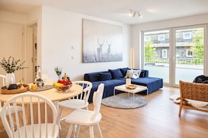 Ein modernes Wohnzimmer mit einem blauen Sofa, weißen Sesseln, einem Esstisch mit Obstschale und einem großen Fenster mit Blick auf Gebäude.