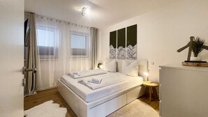 Un dormitorio moderno con una cama grande, una mesita de noche con lámpara, un armario y una ventana con cortinas translúcidas.