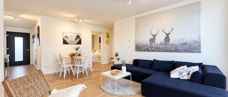 A modern living room with a dark blue sofa, a white chair, a coffee table, and a painting of deer on the wall.