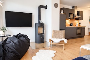 Ein modernes Wohnzimmer mit einem schwarzen Sofa, einem weißen, flauschigen Teppich und einem Holzstuhl. In der Ecke steht ein schwarzer Kaminofen.
