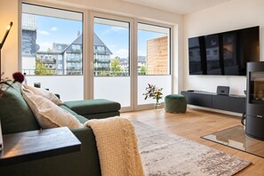 A modern living room with a green sofa, a flat-screen TV, and a kitchen area with a built-in oven. - Moderne Ferienwohnung im Stadtzentrum: 3 Zimmer, Südbalkon und Infrarotkabine für Max. 4 Personen - (Winterberg)