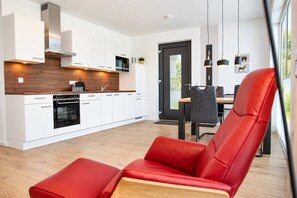 Une cuisine moderne avec des armoires blanches, un four encastré et un plan de travail en bois. On y trouve un coin repas avec une table et des chaises, et une chaise rouge au premier plan.