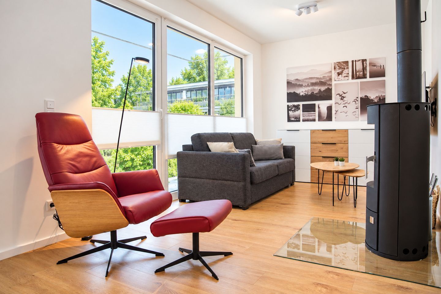 A modern living room with a red chair, grey sofa, and a black stove.