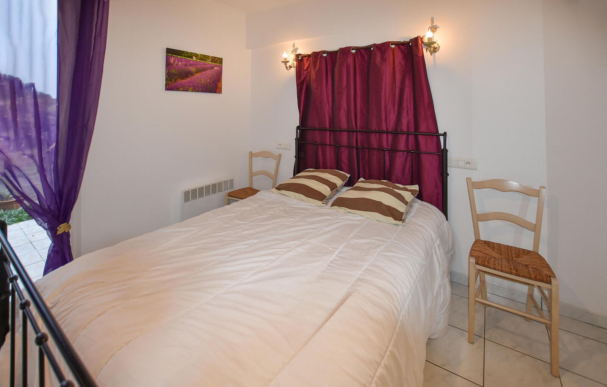 A bedroom with a bed, two chairs, a window with purple curtains, a wall-mounted light, and a small painting.