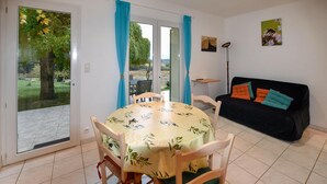 Un área de comedor con una mesa redonda y sillas, un sofá negro con cojines de colores, y vistas a un jardín exterior a través de un ventanal.