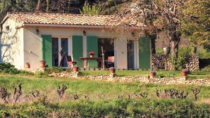Un edificio rústico con techo de tejas, contraventanas verdes y pared de piedra. Hay una mesa y sillas afuera, rodeadas de macetas con plantas y un camino de piedra.