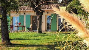 Un edificio rústico con contraventanas verdes, una mesa con sillas y un árbol en primer plano.