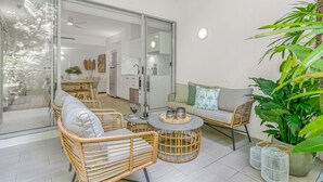 Un espacio de sala moderno con mobiliario de mimbre, una puerta corrediza de vidrio que da a una cocina y una planta grande en maceta.