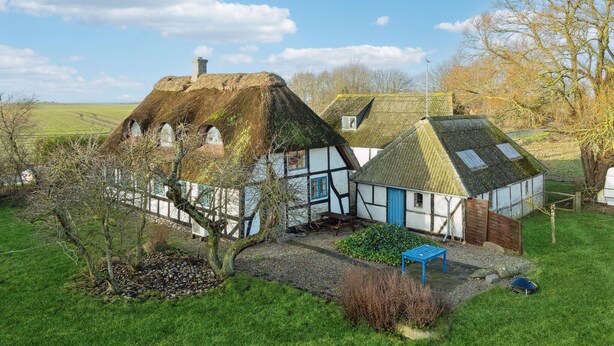 Une maison traditionnelle à colombages, dotée d’un toit de chaume, entourée d’un jardin bien entretenu et d’un paysage rural.