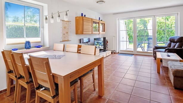 A spacious living area with a wooden dining table and chairs, a sofa, and a TV. There is a sliding glass door leading to an outdoor patio. - Haus 99 - Seepark Burhave by Interhome (Burhave)