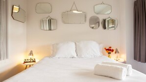 Un letto rifatto con lenzuola bianche e due cuscini, un comodino con una lampada, e un vaso con fiori.