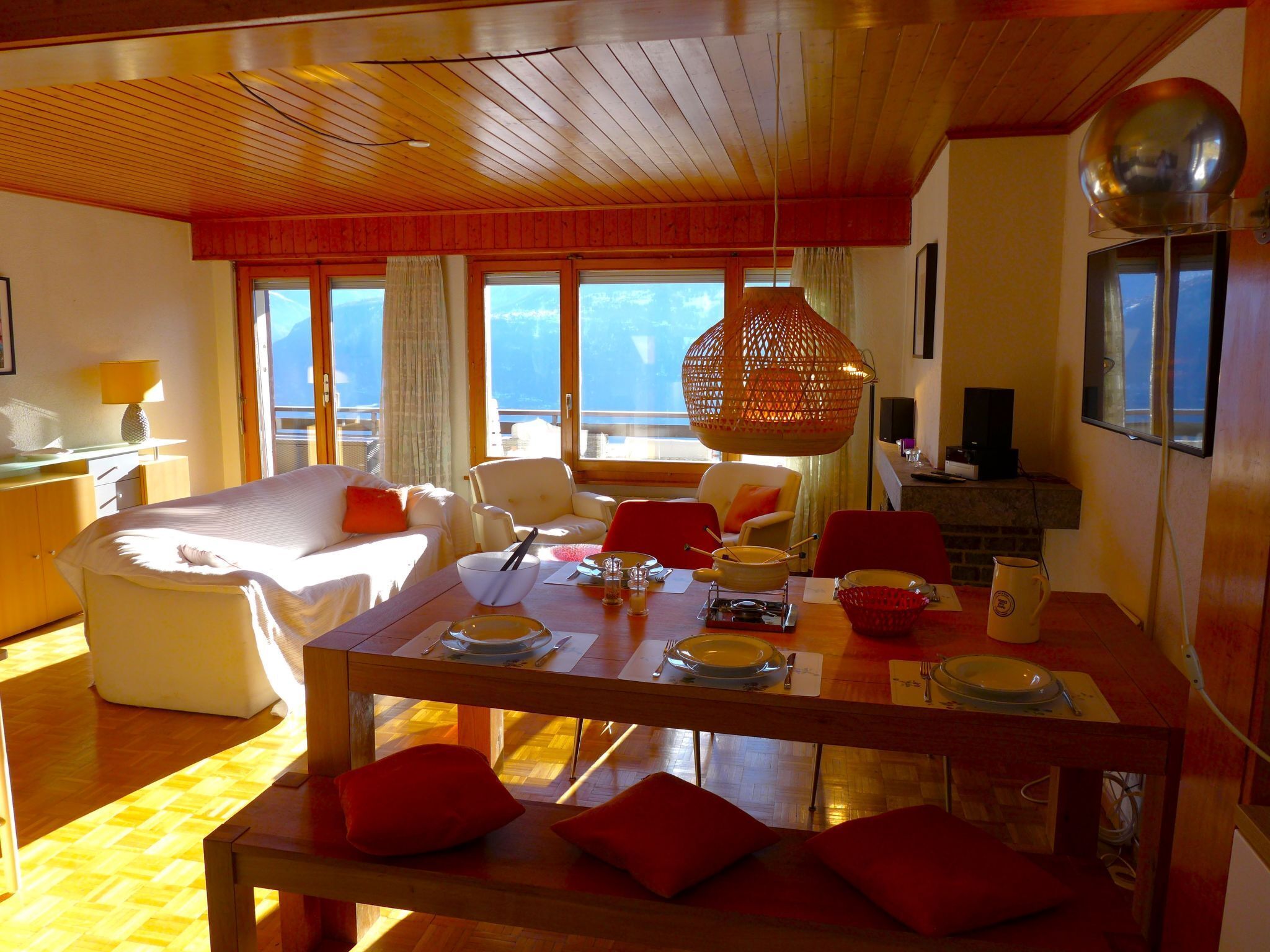 Una sala de estar moderna con techo de madera, una mesa de comedor preparada para cuatro comensales y un amplio ventanal con vistas al exterior.