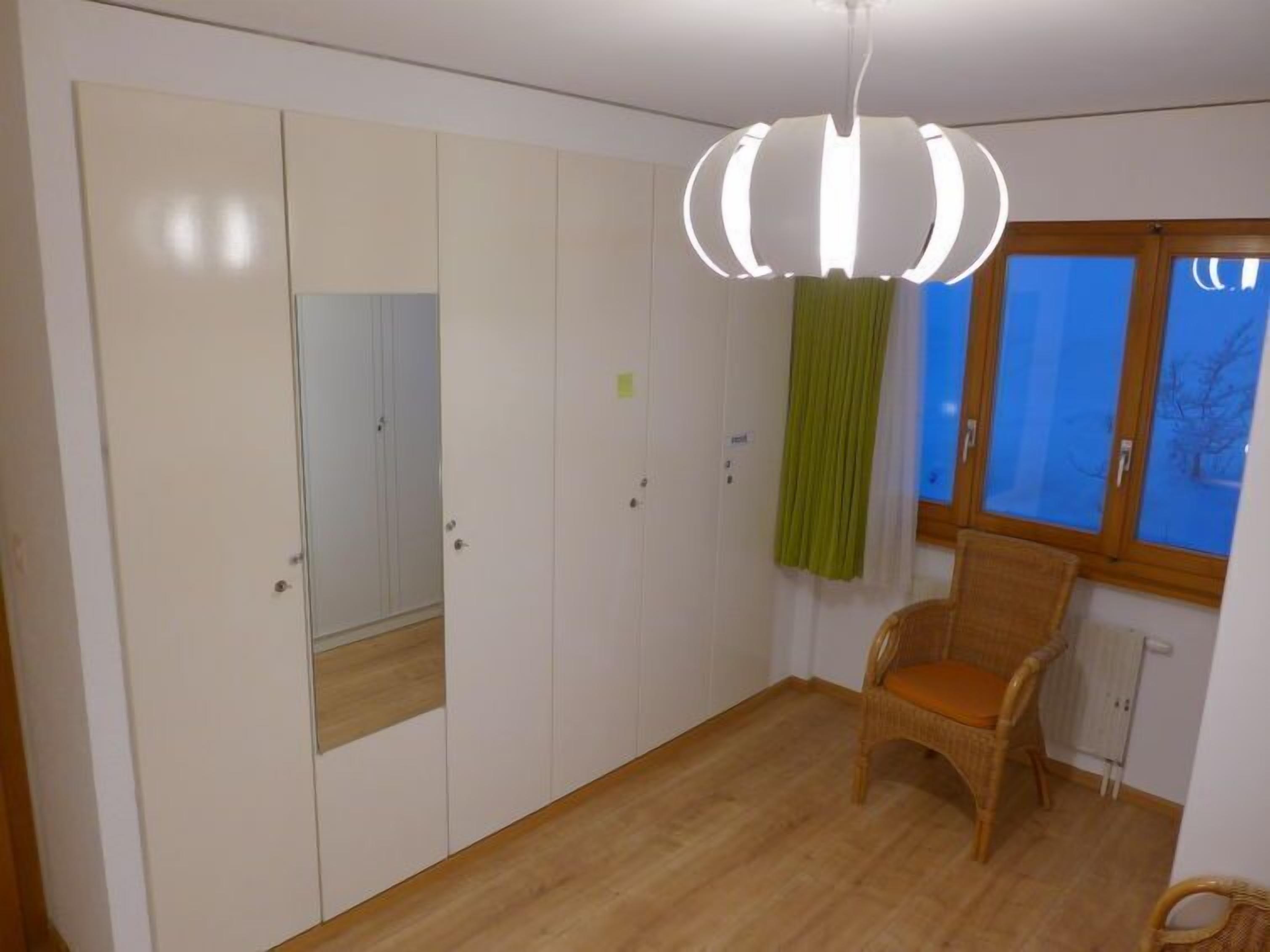 A room with a wardrobe, a chair, a window with curtains, and a hanging light fixture.