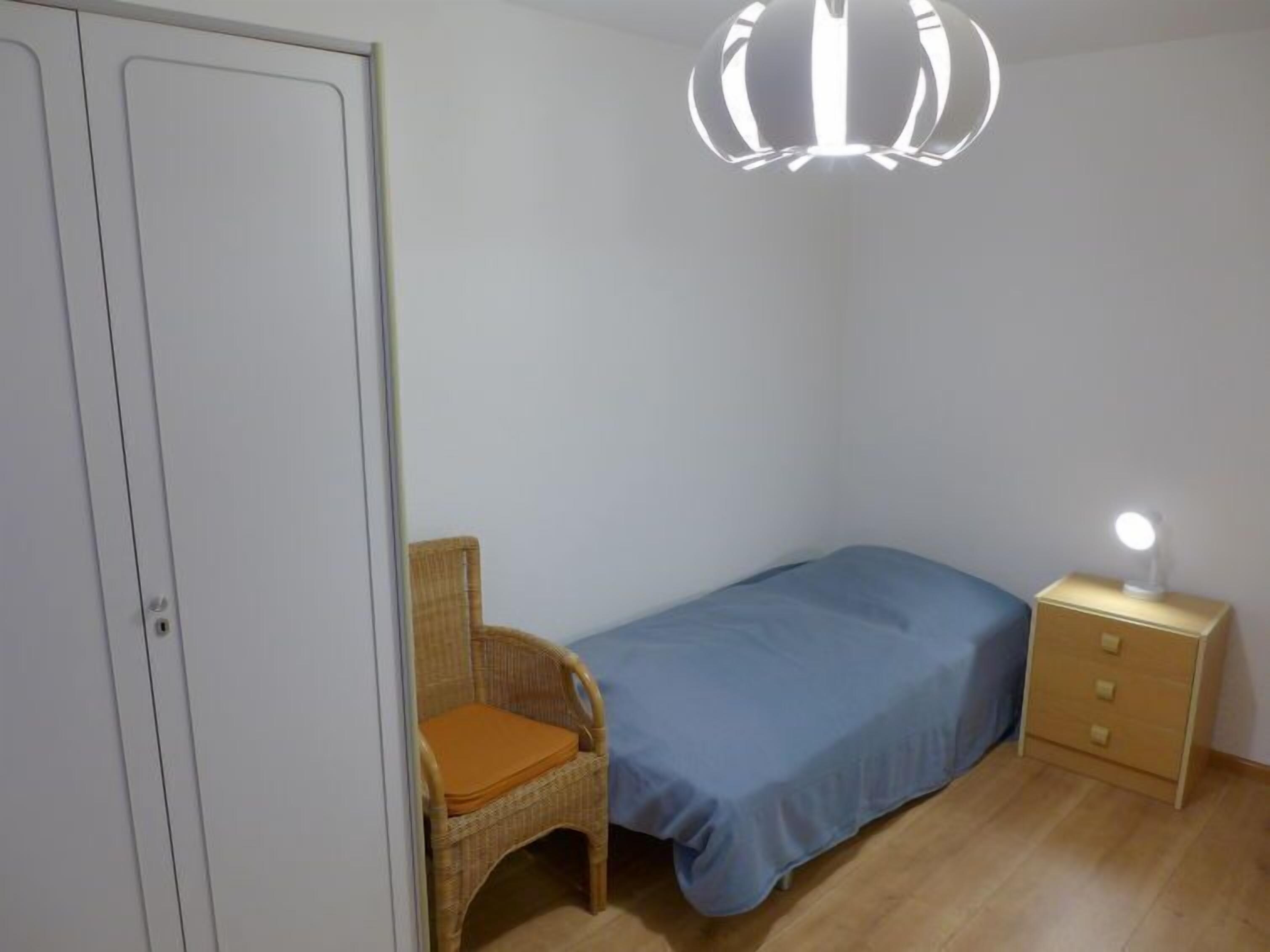 Un dormitorio pequeño con una cama individual, una mesita de noche con lámpara, una silla de mimbre y un armario.