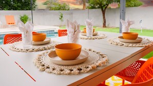 Mesa de comedor dispuesta con cuencos anaranjados y servilletas blancas, rodeada de sillas anaranjadas, con una piscina y follaje al fondo.