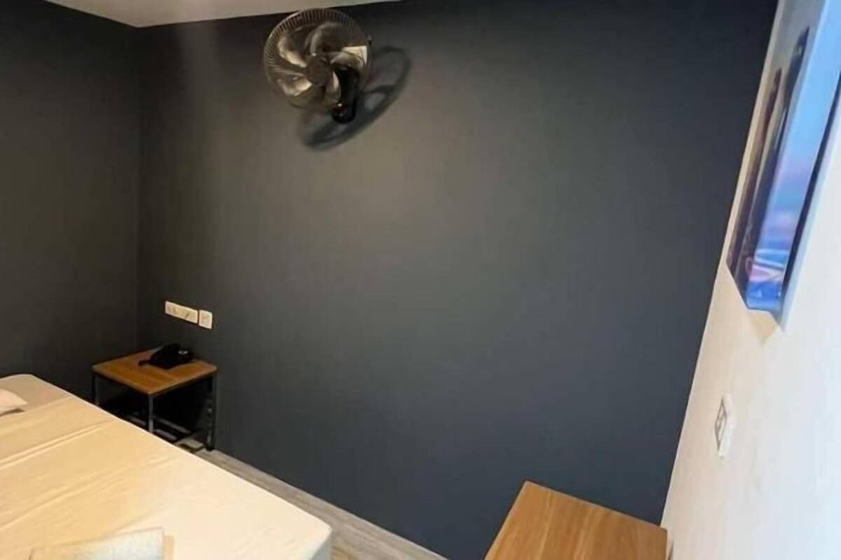 Een kamer met een bed, een klein tafeltje, een ventilator aan de muur en een stopcontact.