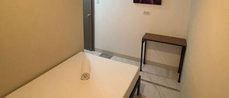 A small, simple room with a bed, a small table, and a wall-mounted shelf.