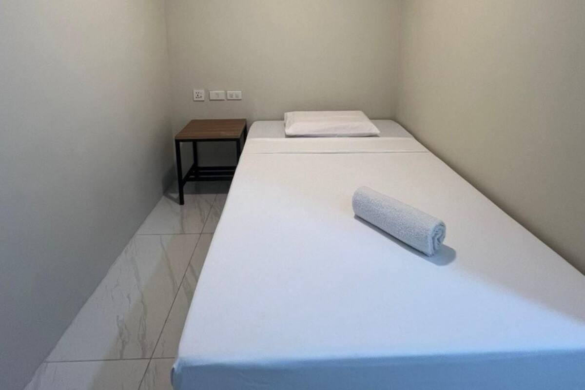 Een kleine kamer met een enkel bed, een houten nachtkastje en een witte spreidek.