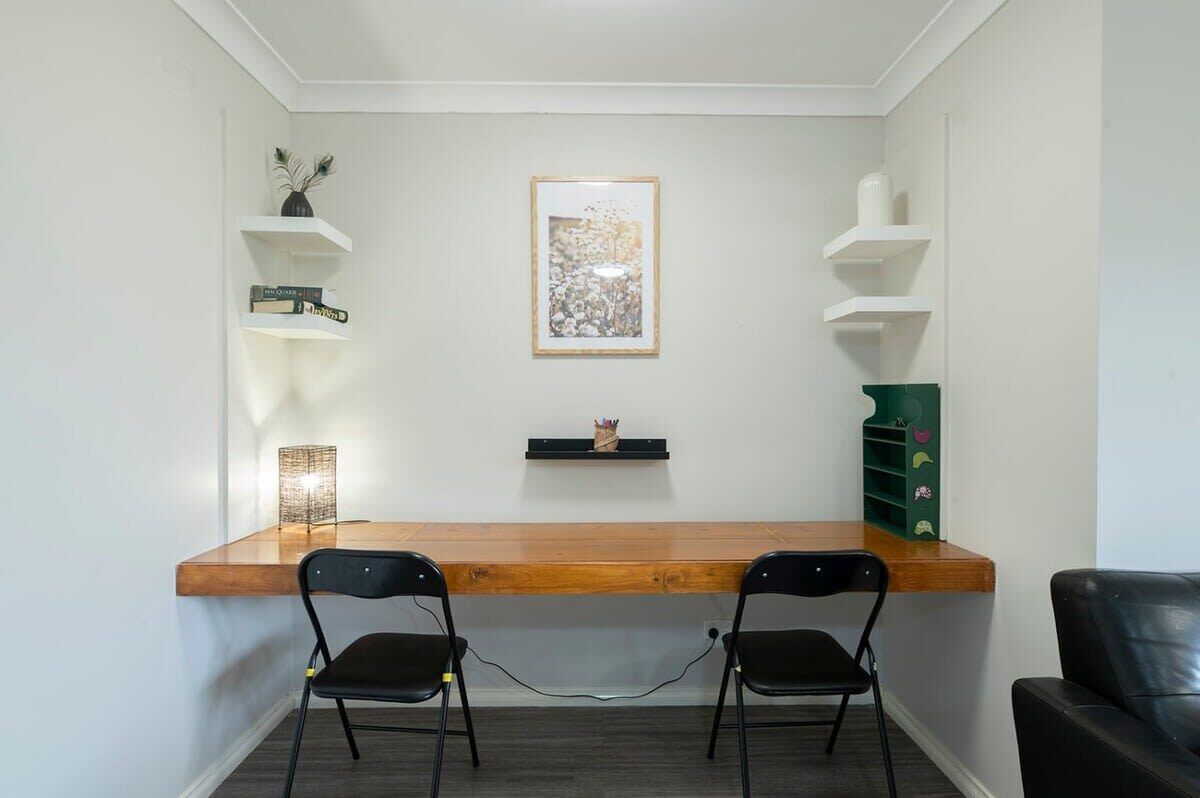 Een kleine, moderne kamer met een houten bureau en twee zwarte stoelen. De muren zijn wit, er hangt een ingelijst kunstwerk aan de wand en er staan planken met diverse spullen.