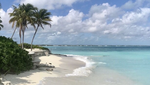 Ein Strand mit klarem, türkisblauem Wasser, weißem Sand und Palmen unter blauem Himmel mit Wolken.