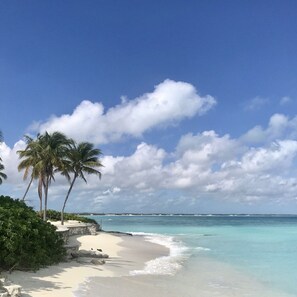 Una playa con agua turquesa cristalina, arena blanca y palmeras bajo un cielo azul con nubes.